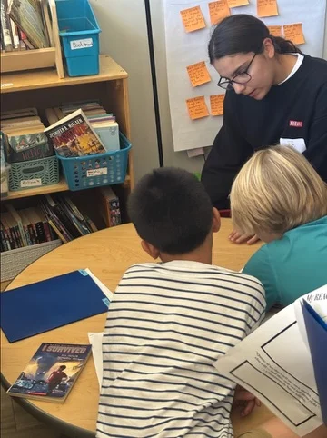 A student instructor working with two younger students around a classroom table.