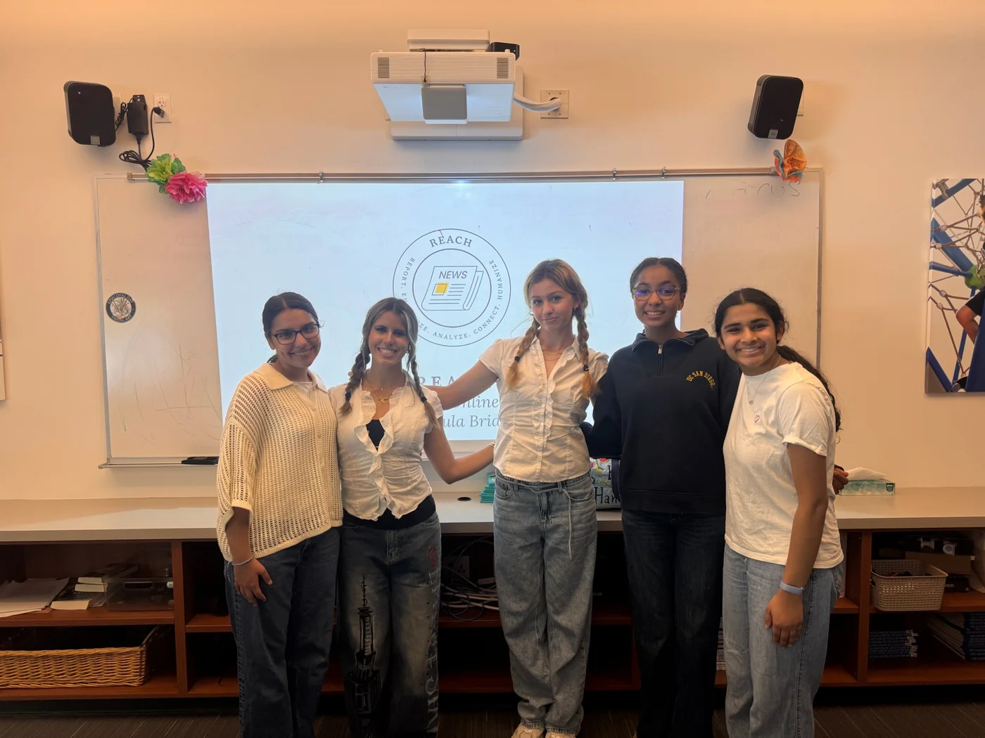 Five REACH team members standing together in front of a classroom screen displaying the REACH logo.